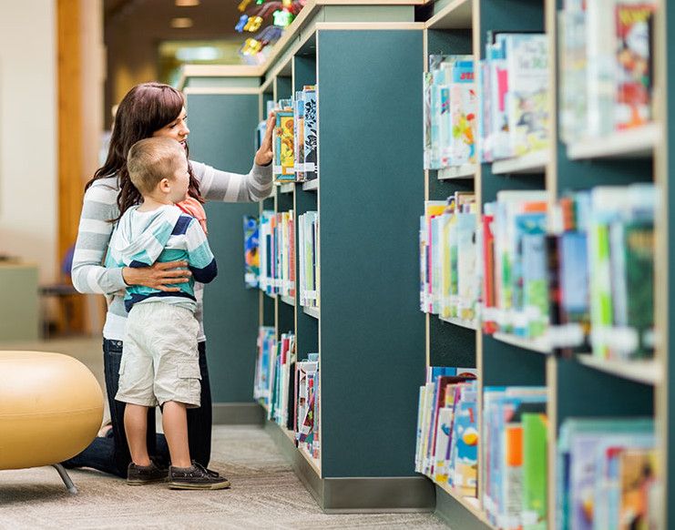 Frau mit Kleinkind, die ein Buch aus einem Bücherregal in der Bibliothek auswählen