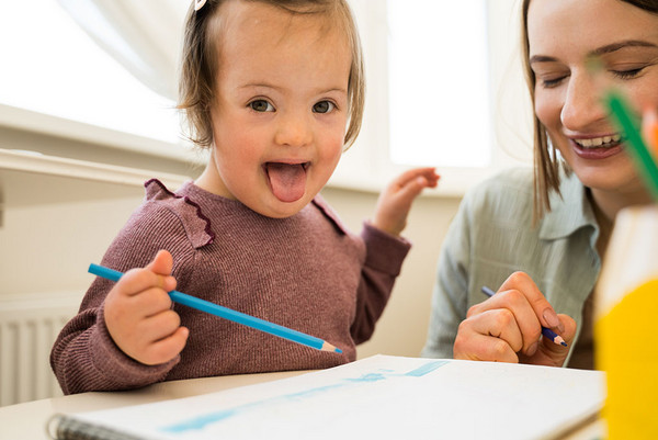 Kleines Mädchen mit Down-Syndrom sitzt an einem Tisch und malt mit ihrer Mutter