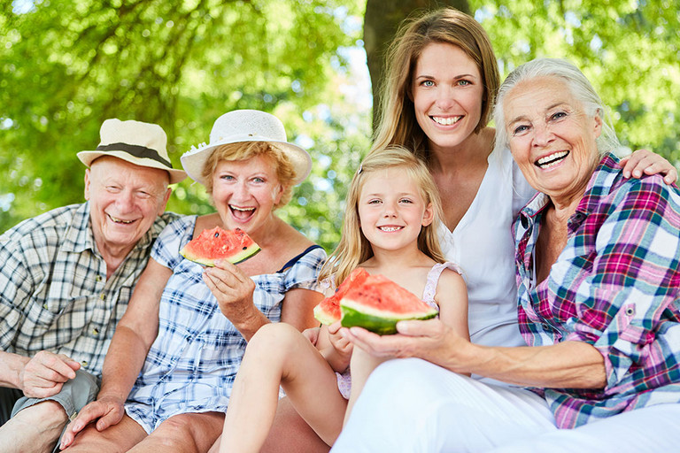 Lachende Gro&szlig;familie mit Gro&szlig;eltern und Kind im Park im Sommer beim Melone essen
