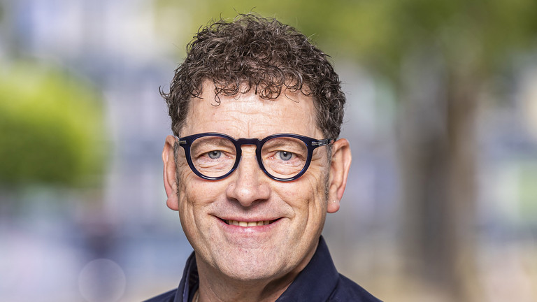 Das Foto zeigt das Portrait eines mittelalten Mann mit dunklen, kurzen lockigen haaren, einer dunklen Brille. Er tr&auml;gt ein dunkelblaues Hemd.