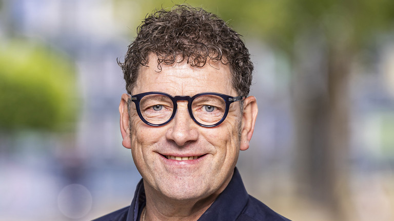 Das Foto zeigt das Portrait eines mittelalten Mann mit dunklen, kurzen lockigen haaren, einer dunklen Brille. Er tr&auml;gt ein dunkelblaues Hemd.
