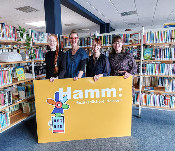 Zu sehen sind vier Frauen, Mitarbeiterinnen der Bezirksbücherei Heessen. Sie halten ein gelbes Schild mit dem Logo der Stadtbüchereien Hamm vor sich fest. Sie lächeln und blicken in die Kamera. Im Hintergrund ist der Innenraum der Bezirksbücherei Heessen zu sehen, die Frauen stehen vor zwei Bücherregalen.
