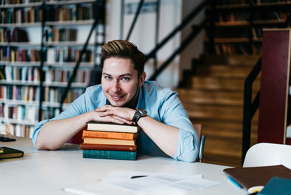 AdobeStock 362857175 Ein junger Mann legt in einer Bibliothek sein Kinn auf einen Stapel Bücher