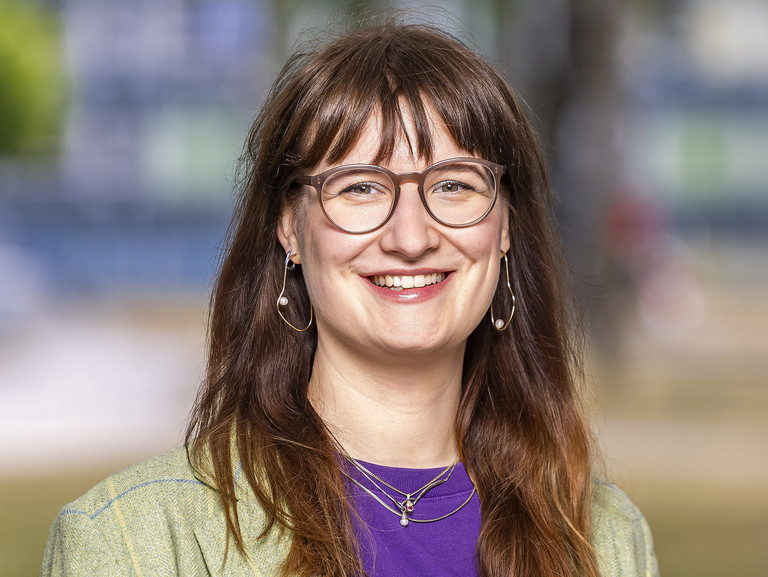 Das Foto ist ein Portrait einer jungen Frau mit dunkelblonden. offenen Haaren, die in die Kamera l&auml;chelt. Sie tr&auml;gt ein lila Shirt und eine hellgr&uuml;ne Jacke.
