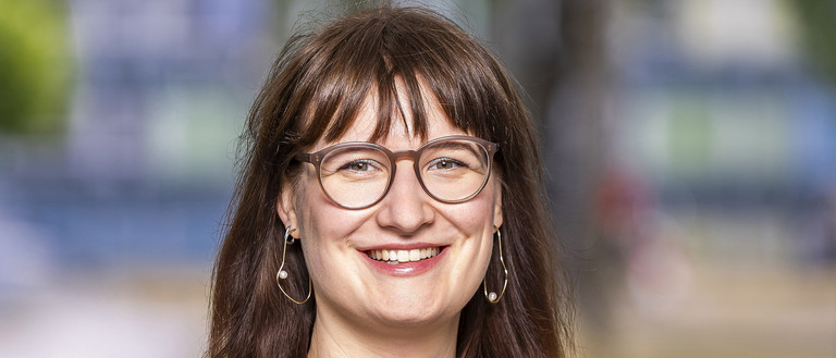 Das Foto ist ein Portrait einer jungen Frau mit dunkelblonden. offenen Haaren, die in die Kamera l&auml;chelt. Sie tr&auml;gt ein lila Shirt und eine hellgr&uuml;ne Jacke.