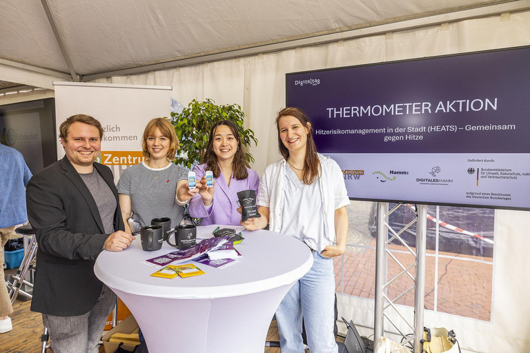 Mitglieder des Verbundprojekts HEATS beim Digitaltag am 8. Juni 2024. Das Foto zeigt einen Mann und drei Frauen, die an einem Infostand stehen. Auf dem Bildschirm im Hintergrund steht "Thermometeraktion".