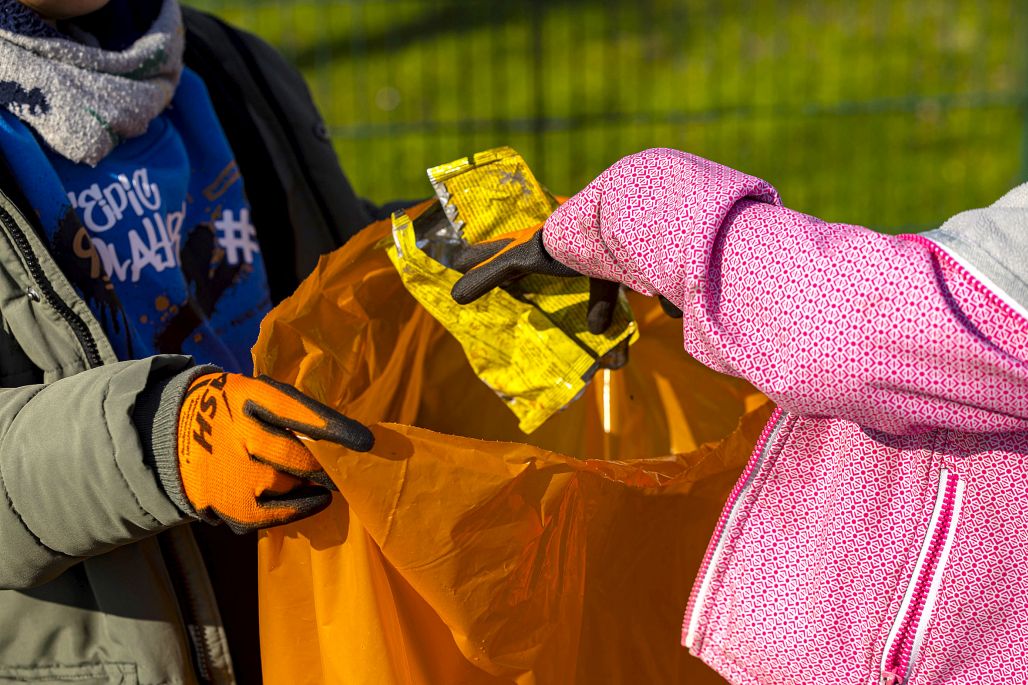 Ein Kind hält einen Müllsack offen, ein anderes Kind wirft eine Plastikverpackung rein.
