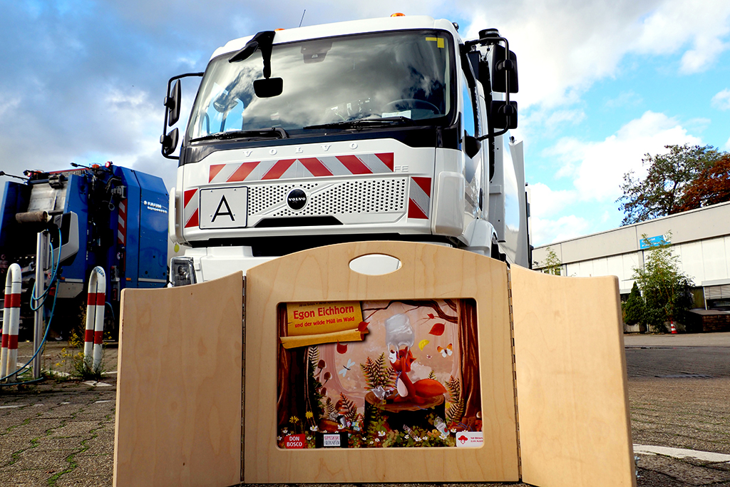 Ein Kamishibai-Theater mit dem Cover der Geschichte "Egon Eichhorn und der wilde Müll im Wald" steht vor einem Müllfahrzeug.
