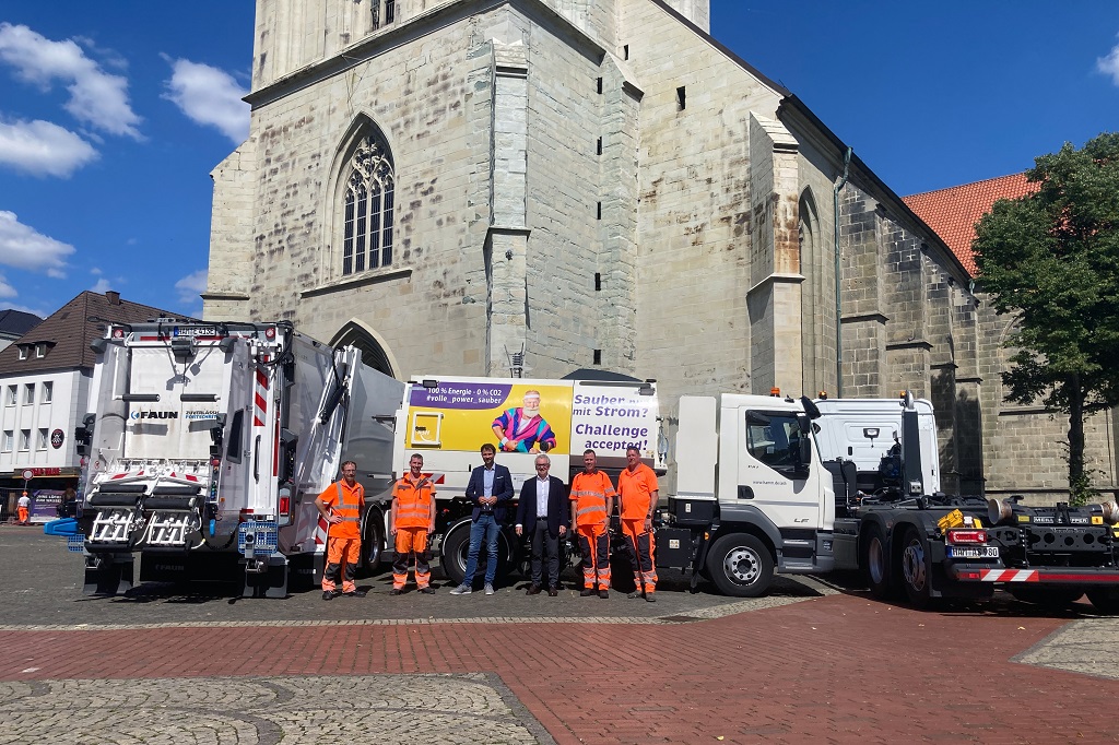 Das Foto zeigt drei Fahrzeuge vor der Pauluskirche in Hamm. Von links nach rechts: Müllfahrzeug, Kehrmaschine, Hakenwagen. Davor stehen Mitarbeitende des ASH.