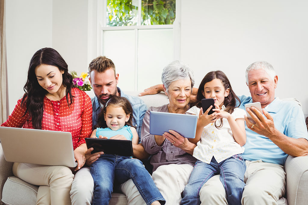 Drei-Generationen-Familie sitzt auf dem Sofa und nutzt Smartphones, Tablets und Laptop