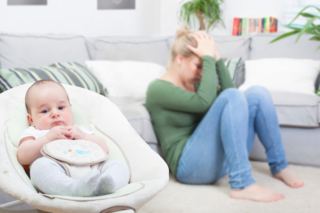 erschöpfte Frau hockt auf dem Boden, Baby sitzt im Vordergrund in einer Wippe erschöpfte Frau hockt auf dem Boden, Baby sitzt im Vordergrund in einer Wippe
