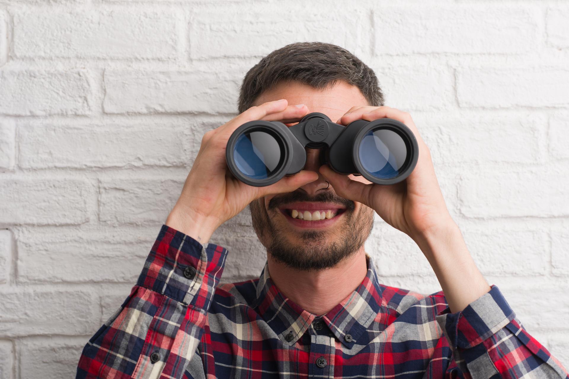 Ein junger Mann, der vor einer weißen Mauer steht und lachend durch ein Fernglas schaut Ein junger Mann, der vor einer weißen Mauer steht und lachend durch ein Fernglas schaut
