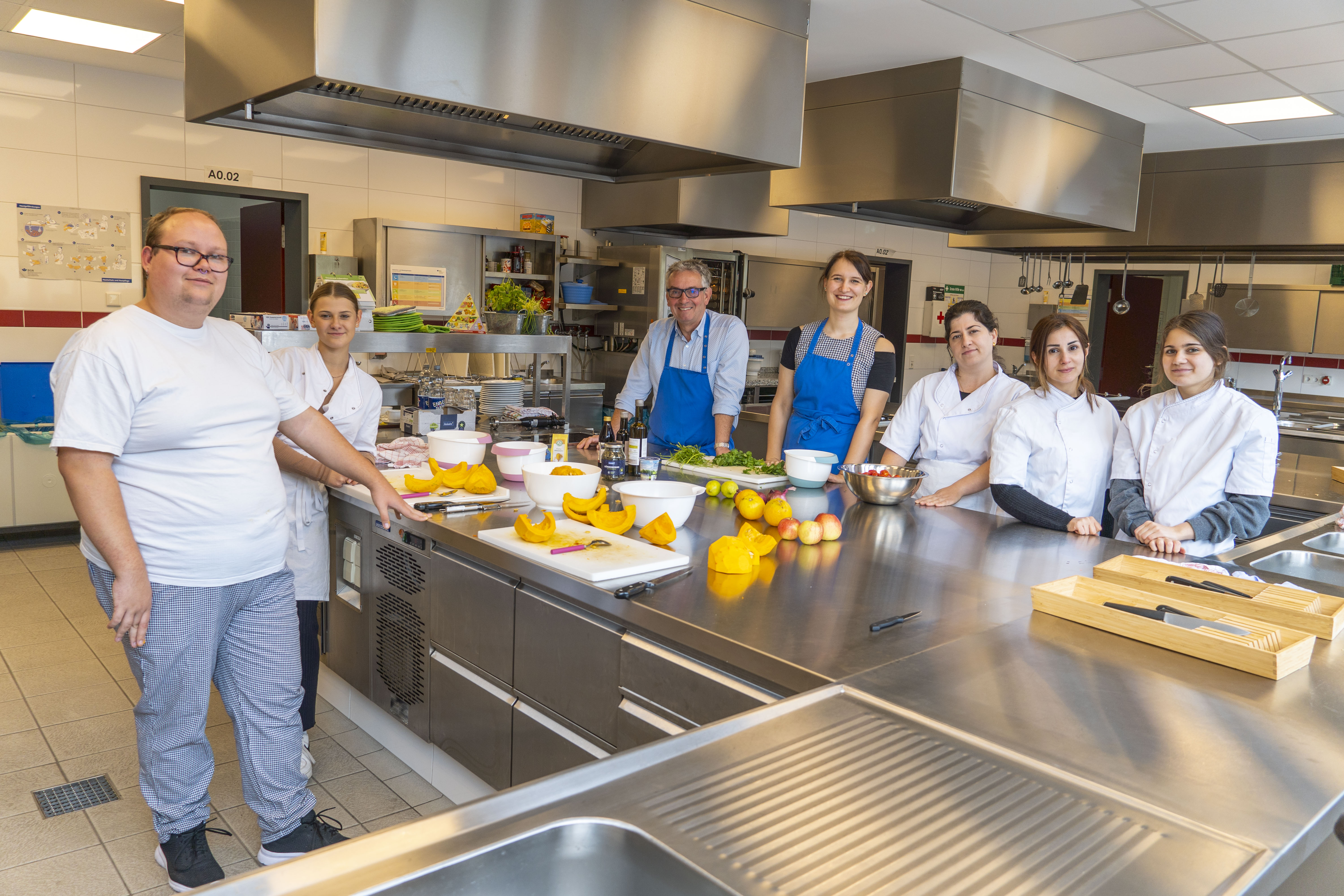 Das Bild zeigt in der Küche des Elisabeth Lüders BK Schüler*innen in Arbeitskleidung, Volker Burgard und Wilma Großmaas mit Schürze in der Küche