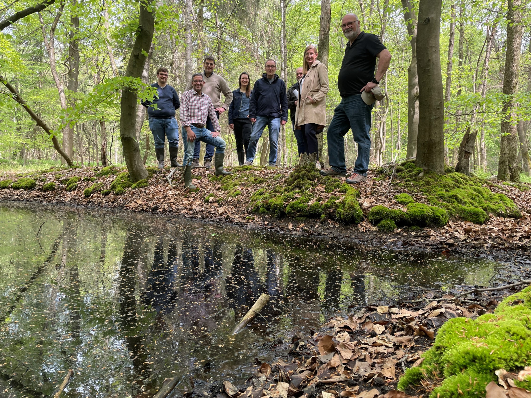 Eine Gruppe von menschen steht vor einem Stillgewässer im frühlingshaften Wald.