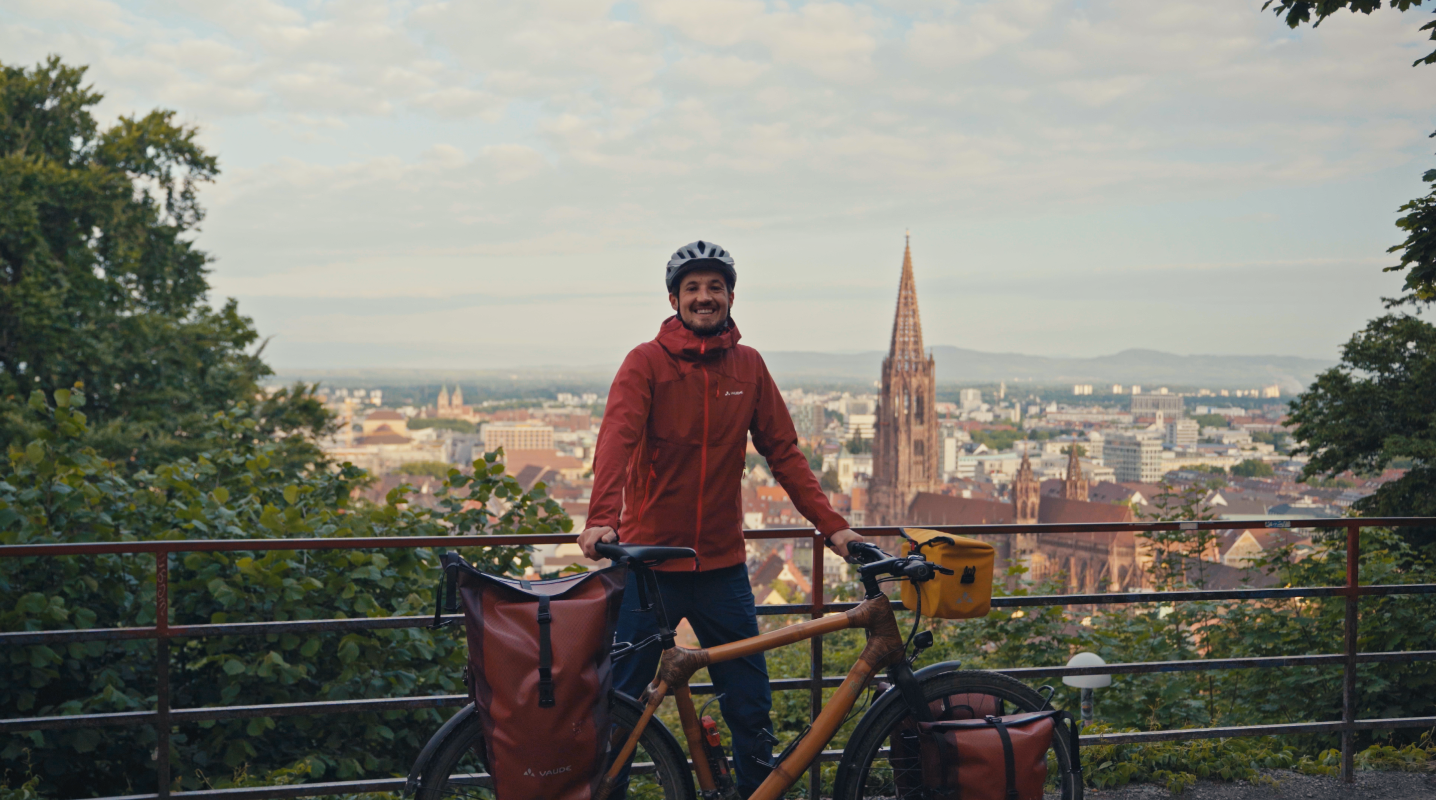 Herr Ingwar Perowanowitsch steht mit seinem Fahrrad auf einer Br&uuml;cke mit der Kulisse von der Stadt Freiburg im Hintergrund.