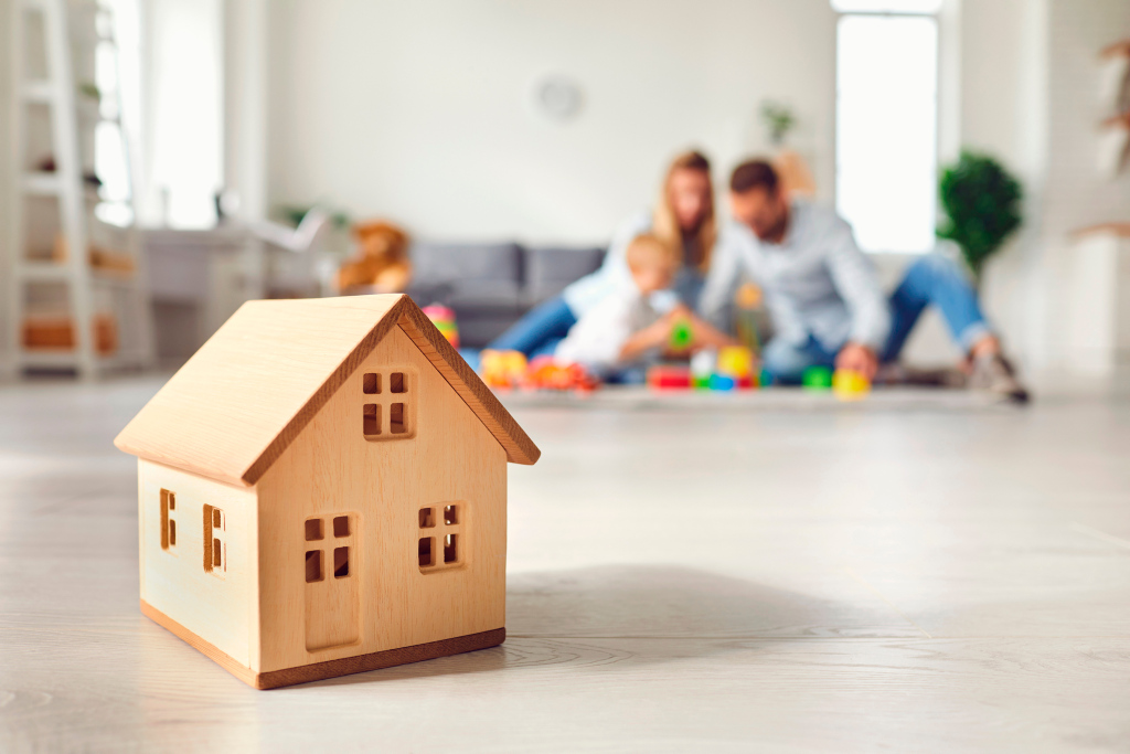 Ein Miniaturholzhaus steht auf einem Fußboden und im Hintergrund spielt eine Familie
