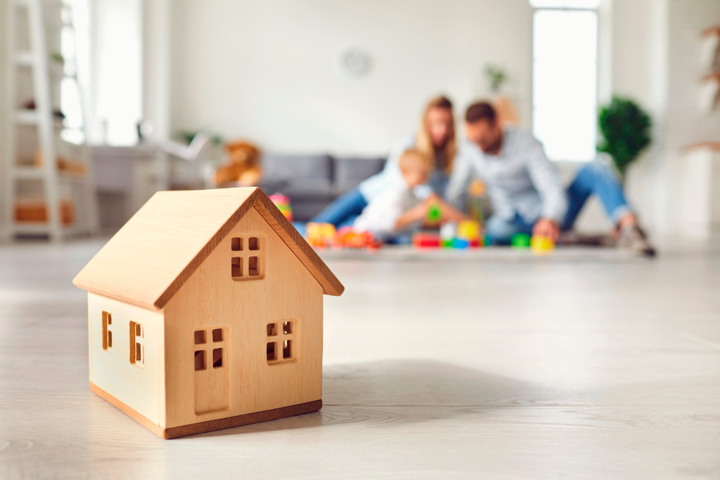 Ein Miniaturholzhaus steht auf einem Fußboden und im Hintergrund spielt eine Familie Ein Miniaturholzhaus steht auf einem Fußboden und im Hintergrund spielt eine Familie