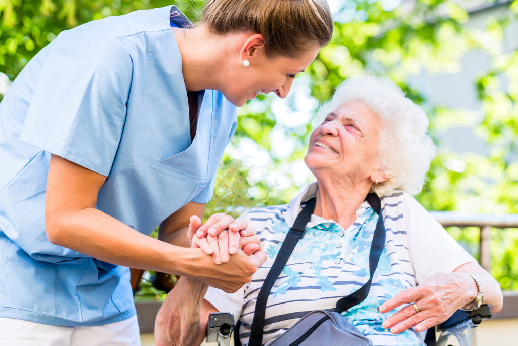 Pflegerin hält Seniorin im Rollstuhl aufmunternd die Hand